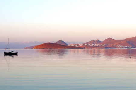 A peaceful sunrise or sunset over a calm, reflective sea, with sailboats and a small island in the distance, bathed in soft pastel light against a backdrop of mountains and a distant coastal town. High quality photoの写真素材