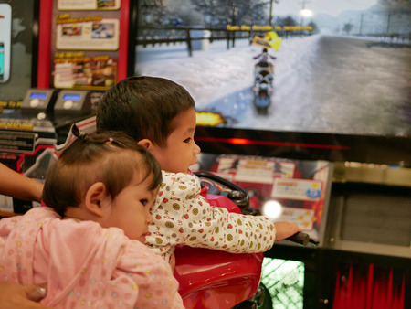 Asian baby, 31 months old, (front) enjoys riding an arcade motorcycle game with her little sister sitting on the backの写真素材