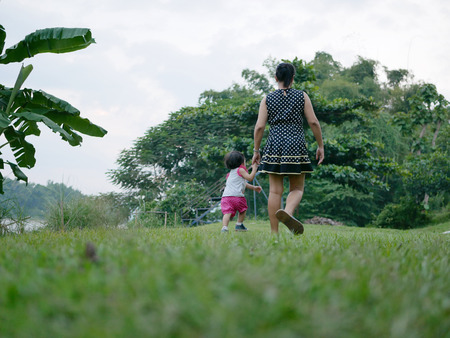 Asian mother holding her little daughter's hand and have a walk together on an outdoor green grass field near a riverの写真素材