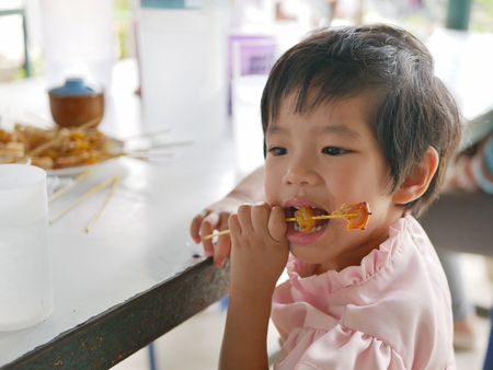 Little Asian baby girl, 34 months old, learning to eat pork satay on a stick by herselfの写真素材