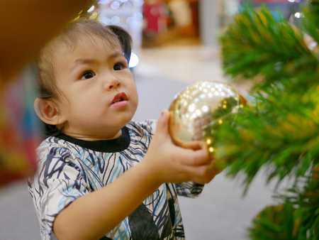 Little Asian baby girl pulling a hanging ball on a Christmas tree in a shopping mallの写真素材