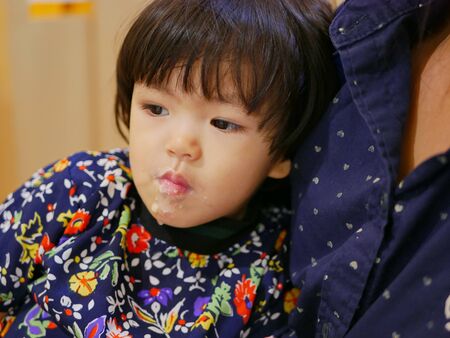 Little Asian baby girl feeling full and leaning on her mother, with her face geting dirty with the condensed milke and ice cream she just ateの写真素材