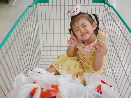 Little Asian baby girl enjoys eating ice cream cone in a cart, while waiting for her mother to do shopping at a supermarketの写真素材