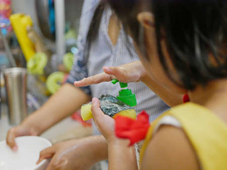 Little baby girl's hand pressing on the cap of a dishwashing liquid bottle, while helping her mother...