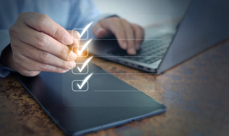 Hand checking digital checklist on tablet with computer, illustrating task organization, corporate efficiency, and productivity strategy for business and office solutions.の写真素材