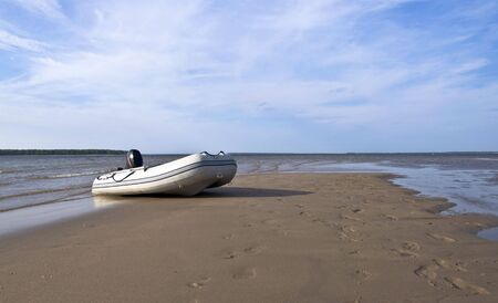 Inflatable fishing boat with an engine standing on a deserted beach on the background of blue sky with clouds. Landscape. の写真素材