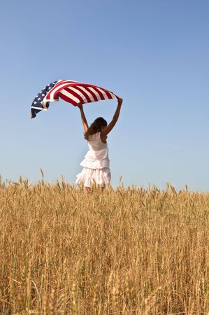 Beautiful young girl holding an American flag in the wind in a field of rye. Summer landscape against the blue sky. Vertical orientation.の写真素材