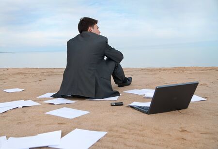 A young businessman sitting on the beach. Around it are scattered documents.の写真素材