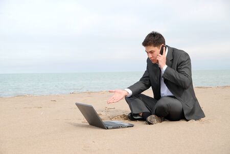 A young businessman sitting c in front of a laptop and talking on a mobile phone.の写真素材