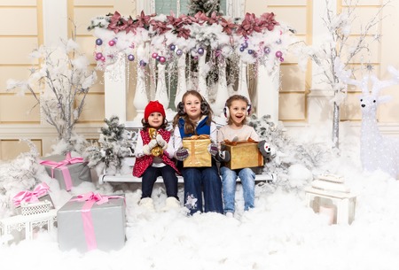Children with Christmas presents sitting on a bench surrounded by toys, gerlyand, gifts, deer and candles. Girlfriends celebrate Christmas together.の写真素材