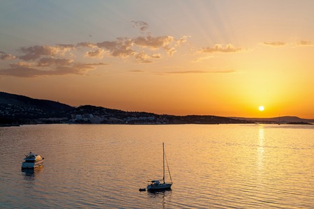 Sunset on the sea. The sun on a bright orange pillow leaving of the day which throwing last rays on the air clouds. In the distance a mountain coast. A yacht and a sailer in the foreground in complete calm.の写真素材