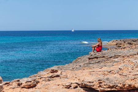 Girl on the beach. Beautiful young girl in red dress sitting on the Spanish coast and looking on the Mediterranean sea.の写真素材
