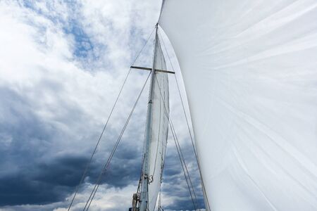 yacht mast with a white sail, the yacht moves with a sail, white sail against a background of cloudy skyの写真素材