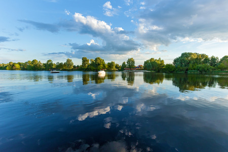 Green trees reflect in the water of the river. In the sky there are dramatic clouds.