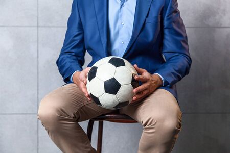 A businessman in a blue jacket sits on a chair and holds a soccer ball in his hands.の写真素材
