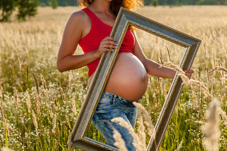 Pregnant girl in nature. Photoshoot of a pregnant girl in jint and red t-shirt in a field at sunset. The girl is photographed with a frame for picturesの写真素材