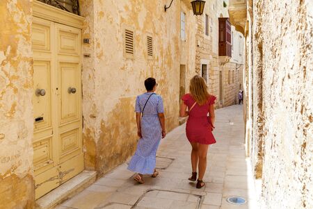 Back view of the women in bright dresses which strolling between the ancient walls of traditional Maltese buildings, Mdina, Malta.の写真素材