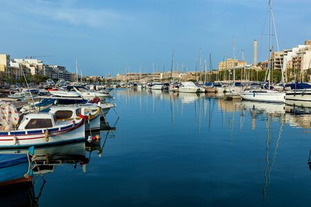 There are a lot of moored yachts in the harbor on the background of Maltese city with old buildings and palm trees on a summer sunny day.の写真素材