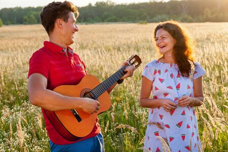 A young man with the guitar is standing near his pregnant wife in the wheat field on a sunny...