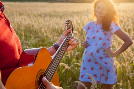 A young man with the guitar is standing near his pregnant wife in the wheat field on a sunny day.Pregnant family photo shoot in natureの写真素材