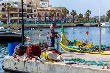 Malta Valletta June 16, 2019: A fisherman is working with fishing nets on the pier near the bright traditional boat on the background of the city view.のeditorial素材
