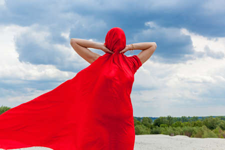A girl in a red cloth on the sand poses for a photographer against the backdrop of a cloudy sky. Red...