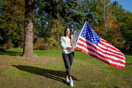 Young beautiful girl with long hair in a white sweater and jeans, laughs and holds a big American...