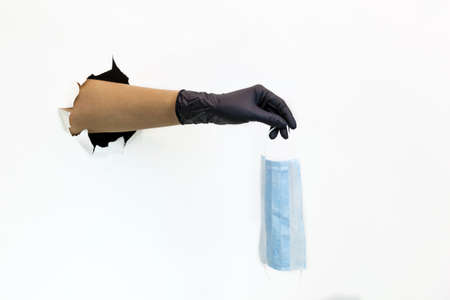 Female hand in a latex black glove in a torn hole on a white background holds a face mask....