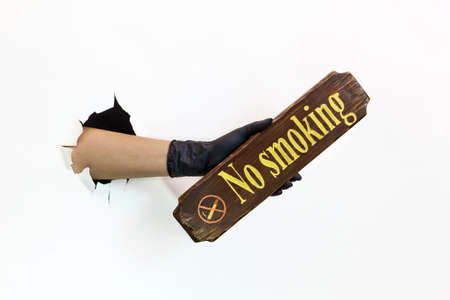 A female hand in a latex black glove in a torn hole on a white background holds a wooden no smoking...
