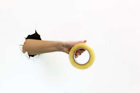 A female hand with a red manicure in a torn hole on a white background holds a roll of tape. Hand...