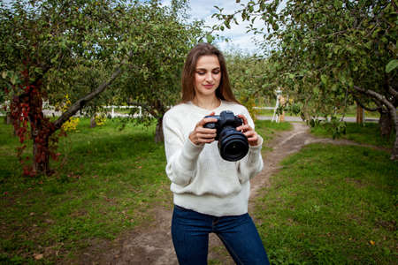 Girl photographer in a white sweater and jeans takes a picture on a professional camera in the park