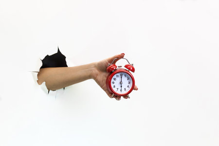 A female hand with red manicure through torn white paper holds a red alarm clock. Hand through torn white paper. Hand holding a red alarm clockの写真素材