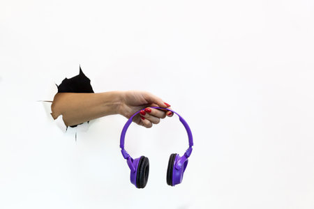 A female hand with a red manicure through torn white paper holds a wireless purple headphone. Hand through torn white paperの写真素材