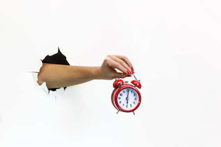 A female hand with red manicure through torn white paper holds a red alarm clock. Hand through torn...