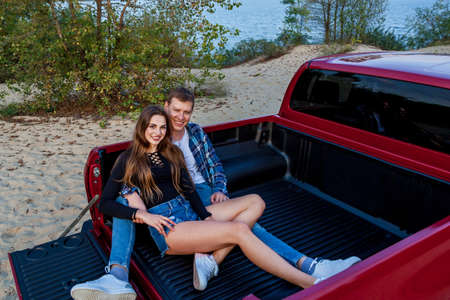 Happy young loving couple smiling and hugging in the back of a red car on the beach near the lake. Young couple hugs near the car.の写真素材