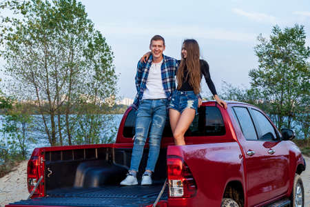 Happy young loving couple smiling and hugging in the back of a red car on the beach near the lake. Young couple hugs near the car.の写真素材