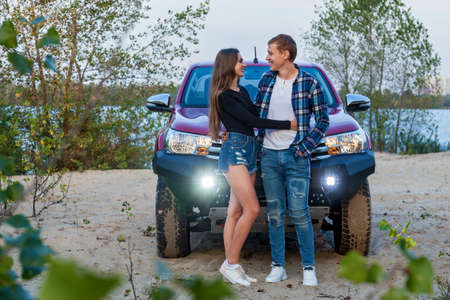 Happy young couple in love smiling and hugging next to red car on the beach near the lake. Young couple hugs near the car.の写真素材