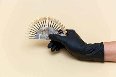 Female hand in a black medical glove holds a coloring scale of artificial teeth for visual measurement of color on a beige background. Concept of dentistry and medical instruments. Photo with place for your text and design.の写真素材