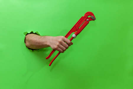 Male hand holds a red pipe key through a torn green paper. Hand through torn green paper. Photo with place for your text, logo and design. Concept repair and toolの写真素材