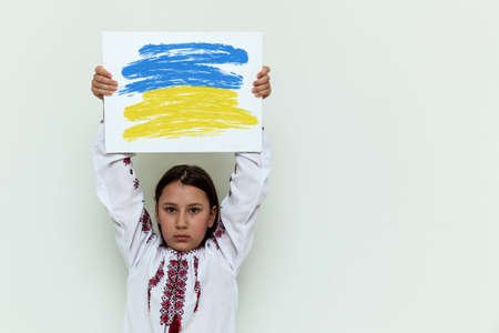 A girl in an embroidered dress stands against the background of a white wall, holding a white sheet with a picture and an inscription. The girl protests against the war.の写真素材