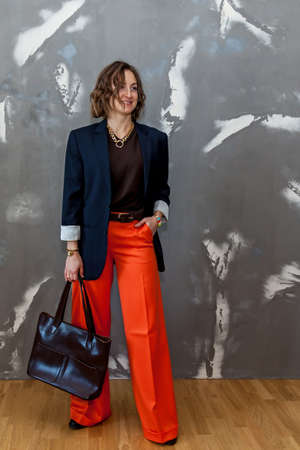 Fashionable and stylish girl posing in orange trousers, a brown sweater and a jacket against a gray wallの写真素材