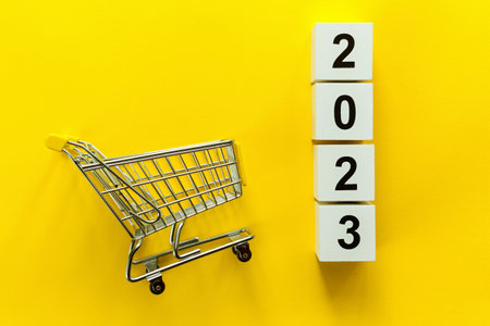 Wooden white cubes with numbers 2023 and a metal trolley on a yellow background. Concept of new year shopping, merry christmas and happy new year.の写真素材