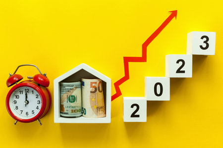 Wooden white cubes with numbers 2023 with a house with money and a red alarm clock on a yellow...