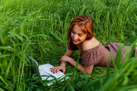 A beautiful girl in a dress lies in a green field of young wheat and works on a laptop, enjoying...