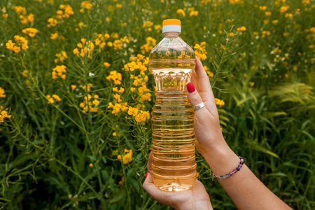 A woman's hand holds a bottle of rapeseed oil against the background of yellow rapeseed flowers