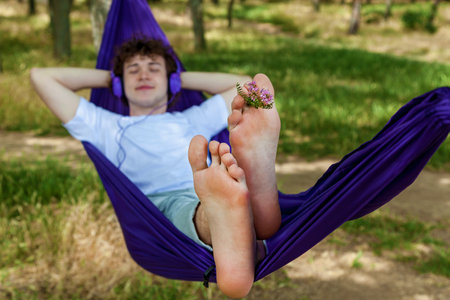A young guy lying in a purple hammock listens to music on headphones while enjoying nature.の写真素材