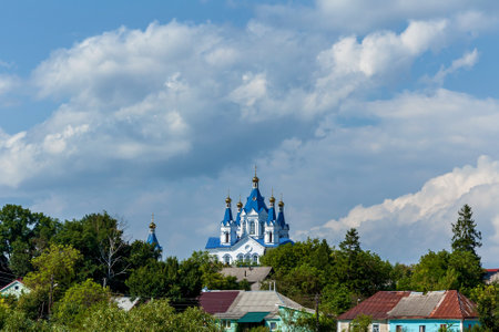 Majestic orthodox church with vivid blue domes and golden crosses stands above a rural village surrounded by green trees, under a dramatic sky with fluffy cloudsの写真素材