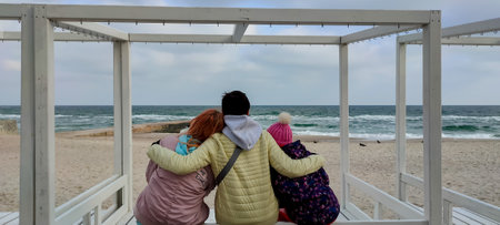 Three people in warm jackets and hats sitting closely on a white wooden bench, embracing and gazing at the waves of a cloudy sea, creating a cozy and emotional winter beach sceneの写真素材