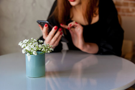 Close-up of delicate white flowers in a small blue vase on a glossy table, with a blurred background of a woman in black dress interacting with her smartphoneの写真素材