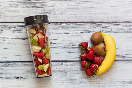Top view of a blender cup filled with sliced bananas, kiwis, and strawberries next to fresh fruits on a weathered white wooden surfaceの写真素材